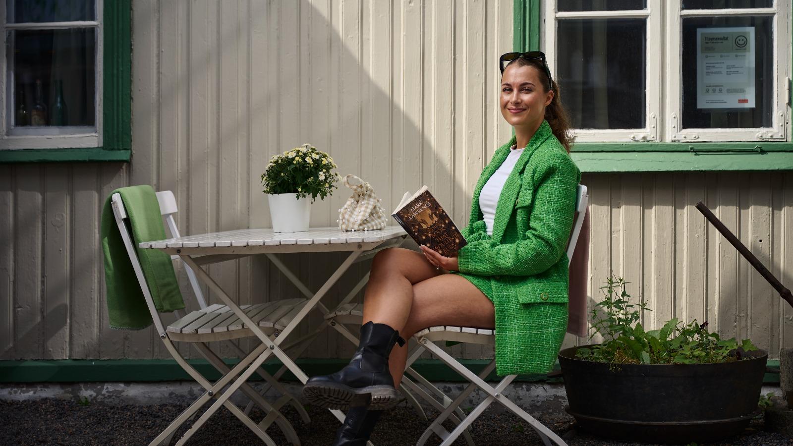 Sophie Verdu sitter ved et cafébord ute og holder en bok mens hun ser mot kamera og smiler